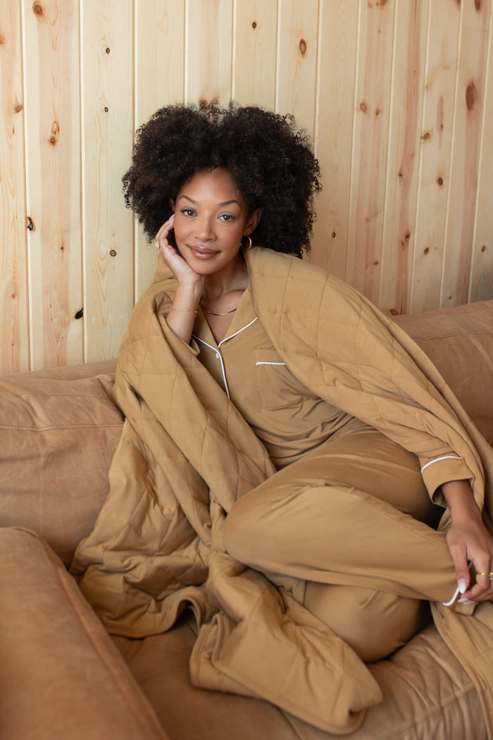 Model sitting on a beige couch covered with Adult Quilted Blanket in Ochre 2.5 wearing Long sleeve women's pajama set in Ochre