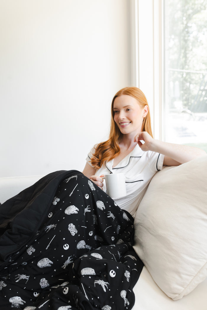 Model sitting on white sofa posing with Adult Quilted Blanket in STAR WARS™ 2.5 print