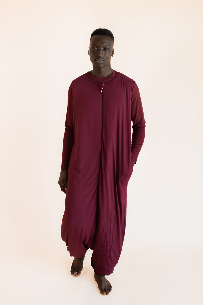 A man wearing a loose-fitting, deep burgundy sleep sack with a front zipper, photographed against a solid light beige background.