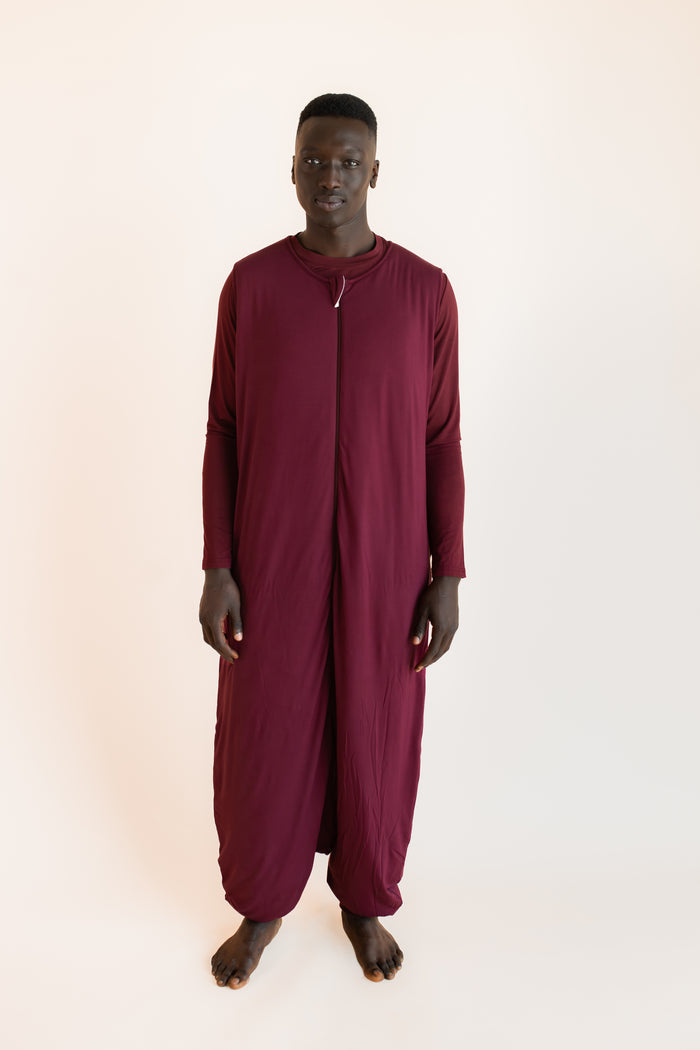A man wearing a full-length maroon sleep sack or caftan-style garment stands against a plain light beige studio background.