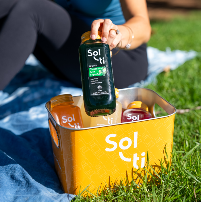 Person holding a Sol-ti SuperAde bottle, sitting on a blue blanket, beside a orange Aluminum Ice Bucket with assorted SuperAde bottles