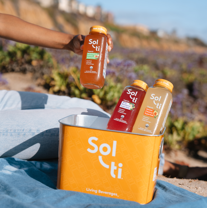 Person holding a Sol-ti SuperAde bottle, sitting on a blue blanket in a flower field, beside a orange Aluminum Ice Bucket with assorted SuperAde bottles
