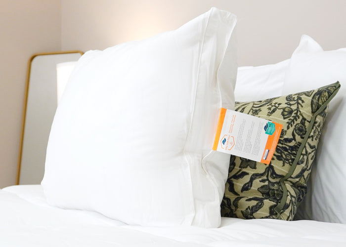 The HD Supply Best Western® Dreammaker Gusseted Pillow with medium-firm support and product tag rests on crisp white bedding, complemented by a green floral pillow and gold-framed mirror for an inviting, hotel-like ambience.