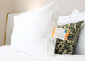 The HD Supply Best Western® Dreammaker Gusseted Pillow with medium-firm support and product tag rests on crisp white bedding, complemented by a green floral pillow and gold-framed mirror for an inviting, hotel-like ambience.