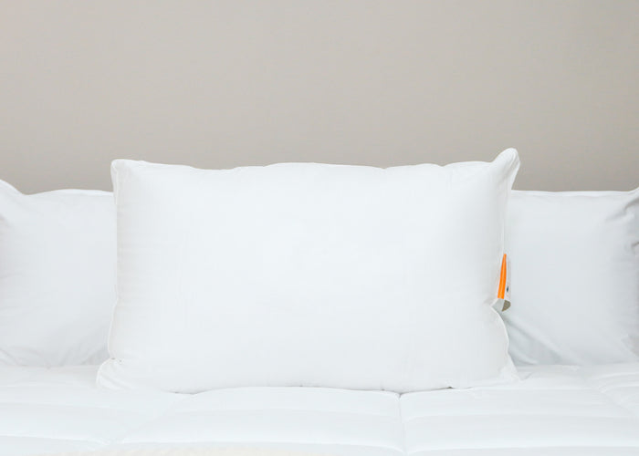 The Best Western® Dreammaker Gusseted Pillow by HD Supply, offering medium-firm support, sits upright on a neatly made bed with a white comforter. A visible orange and gray tag accents the minimalistic setting against a light beige wall.