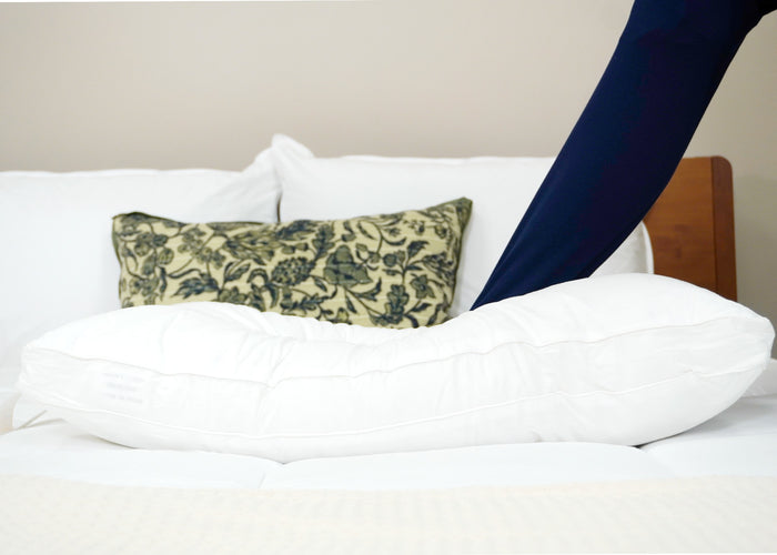 An HD Supply Best Western® Dreammaker Gusseted Pillow with medium-firm support rests on a bed with white sheets. A person in navy blue pants places their leg over the C-shaped pillow, near a green and black floral cushion against the headboard.