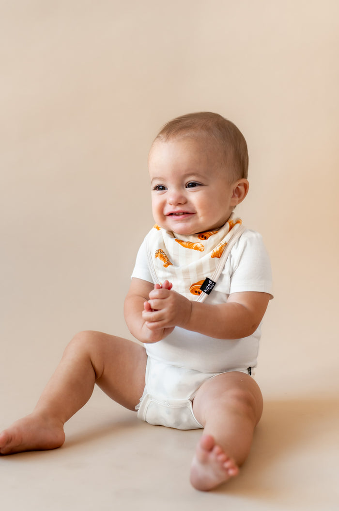 Baby smiling wearing a Bib with Croissant print over white bodysuit on a beige background
