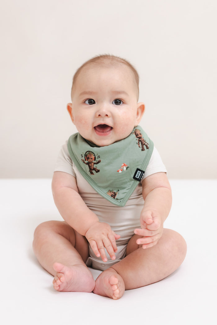 Baby sitting on a beige background wearing Green Bib in Groot print over a beige bodysuit
