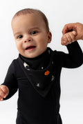 Baby wearing Bib in Halloween Friends print over a black romper holding a hand on a white background