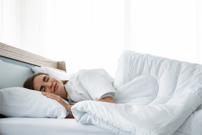 A person is peacefully sleeping in bed, enjoying luxurious comfort. They are lying on their side with a Blue Ridge TriLoft™ Hotel Chamber Pillow, which is suitable for all sleep positions and offers a hypoallergenic experience, under their head and a thick, white quilt covering them. The room is well-lit with a soft, calm ambiance, and sheer white curtains allow natural light to filter in.