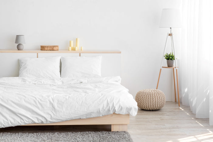 A minimalist bedroom showcases a wooden bed adorned with white bedding and the Blue Ridge TriLoft™ Hotel Chamber Pillow, presenting luxurious comfort for all sleep positions. Two pillows lean against a light wood headboard. A stand featuring candles, a table lamp, and a decorative basket enhances the decor. Natural light filters through sheer curtains beside the tripod floor lamp and small plant.