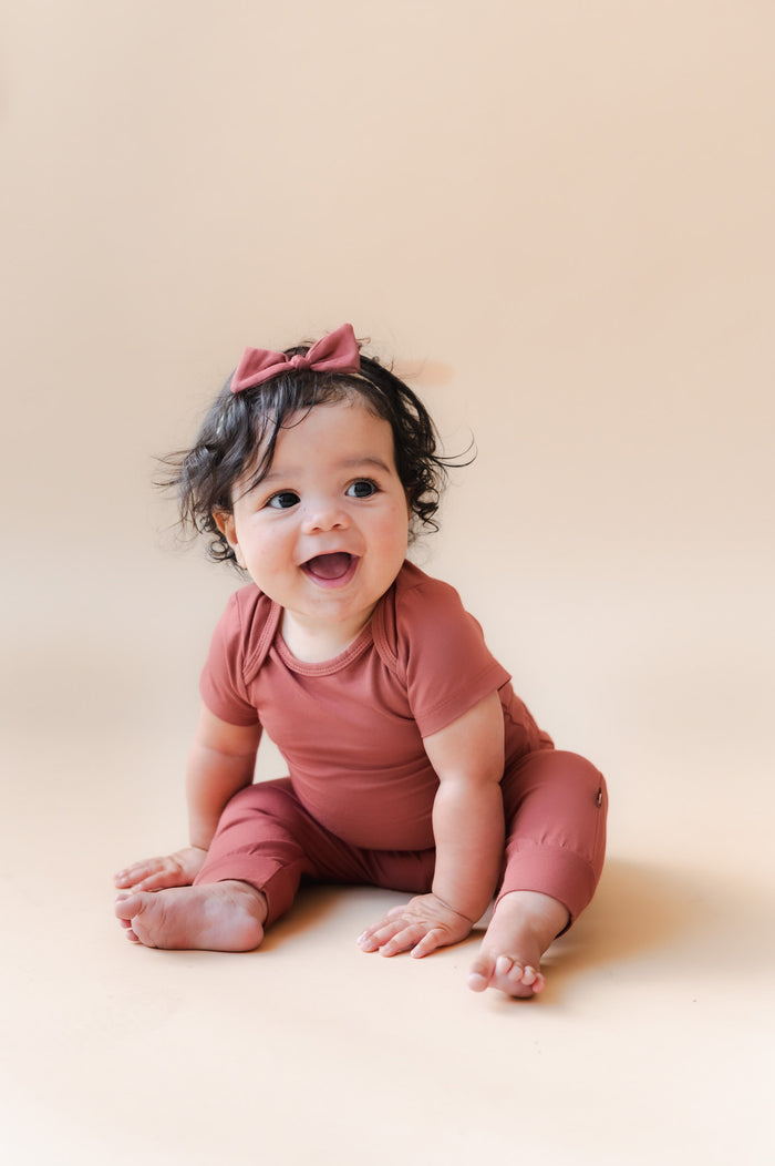 A smiling baby sitting on a warm beige studio background, wearing a matching dusty terracotta short-sleeve bodysuit and matching bow headband.