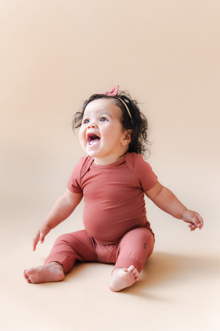 A smiling baby sitting on a warm beige studio background, wearing a matching dusty terracotta short-sleeve bodysuit and a small pink bow headband.