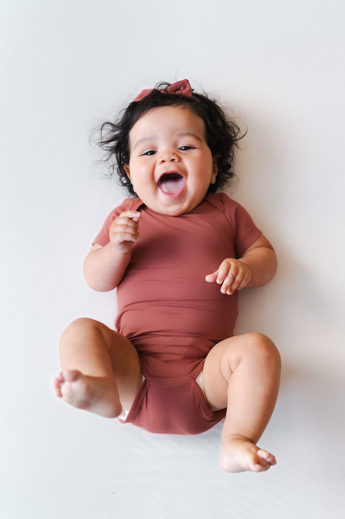 A smiling baby lying on their back wearing a dusty terracotta one-piece bodysuit and a matching hair bow