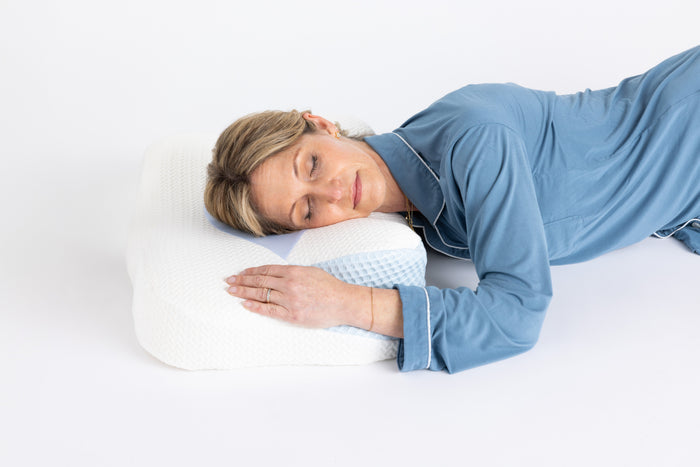 A woman in blue pajamas sleeps peacefully on her side with her arm resting on the Sleep Again Pillows Cervical Memory Foam Pillow for Neck & Shoulder Support, featuring an ergonomic orthopedic design and a textured cover with a blue checkered panel.