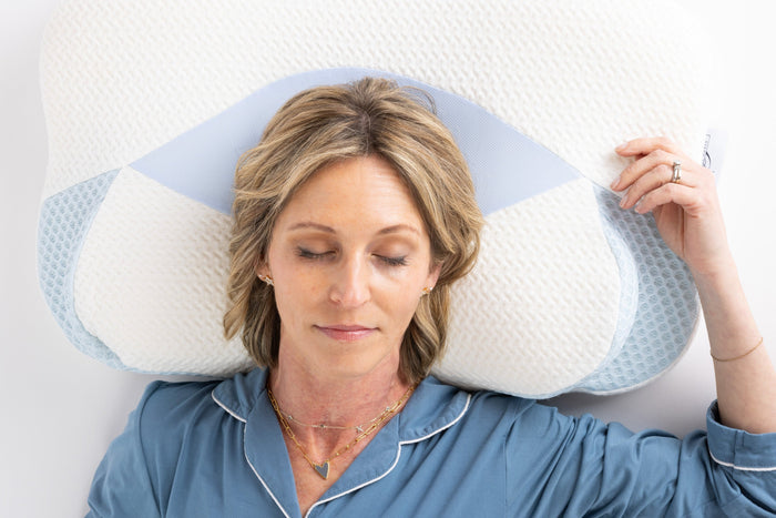 A woman rests with eyes closed, head supported by the Sleep Again Pillows Cervical Memory Foam Pillow for Neck & Shoulder Support. She appears relaxed in blue pajamas, accessorized with layered gold necklaces and rings.