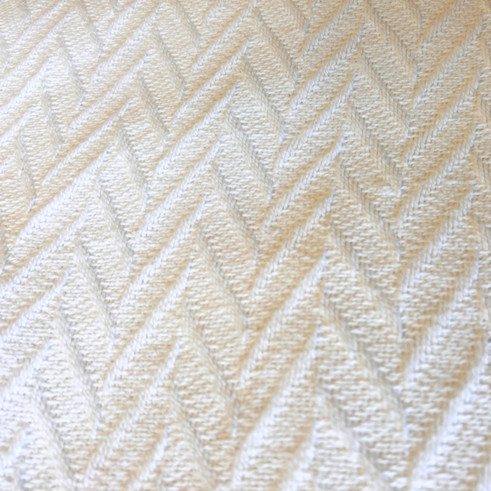 Close-up of a beige textured fabric from the 1888 Mills Cotton Thermal Blanket, featuring a herringbone pattern that evokes luxury. The zigzag design introduces a subtle geometric effect with alternating raised and recessed lines, adding depth to the soft, 100% cotton woven material. Gentle light reflections accentuate its intricate details.