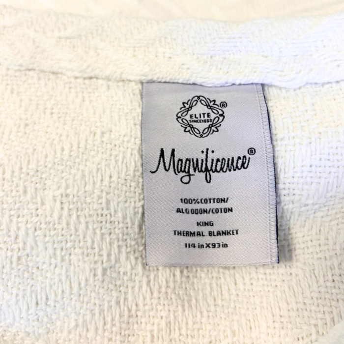 A close-up of a luxurious white textured fabric showcases a label that reads: "1888 Mills, Cotton Thermal Blanket, 100% Cotton, King Size, 114 in x 93 in." The label is sewn onto the premium cotton fabric, which appears soft and intricately woven for five-star hospitality.