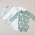 Three baby bodysuits on a light pink background, including a green one with goose pattern and two plain ones.