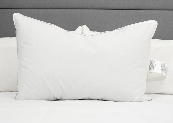The Pillow Factory Downlite 50/50 Waterfowl Feather/Grey Down Pillow stands upright on a neatly made bed with a white quilt and gray upholstered headboard. Another medium support pillow is partially visible behind it.