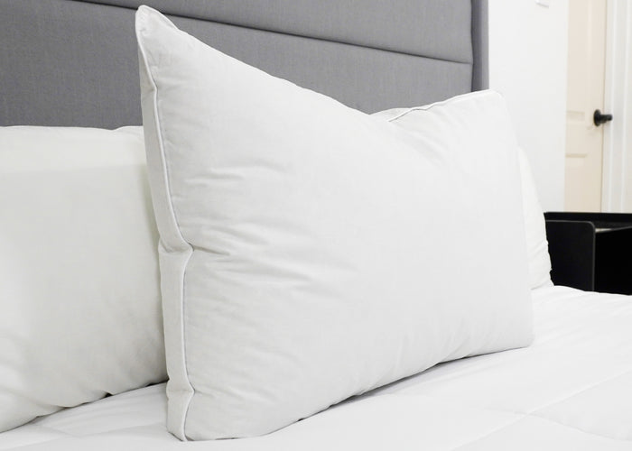 A Downlite 50/50 Waterfowl Feather/Grey Down Pillow by Pillow Factory rests against a gray upholstered headboard on a neatly made bed with white bedding, giving the room a clean, minimalistic look. A white door and dark furniture are also visible.