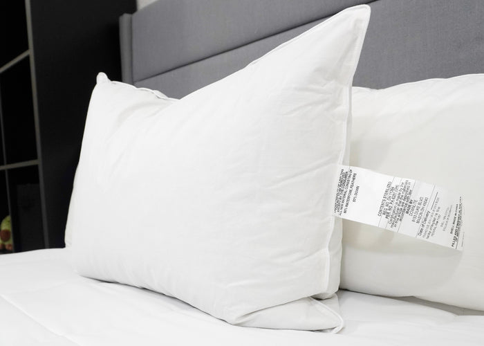 A Pillow Factory Downlite 50/50 Waterfowl Feather/Grey Down Pillow with a care tag rests in front of another medium support pillow on a neatly made bed with white sheets and a gray headboard, shelving partially visible in the modern room.