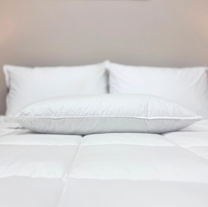 A Downlite 50/50 Waterfowl Feather/Grey Down Pillow by Pillow Factory offers medium support and lies on a neatly made bed with a white comforter, surrounded by three white pillows against a beige wall for a cozy, minimalist look.