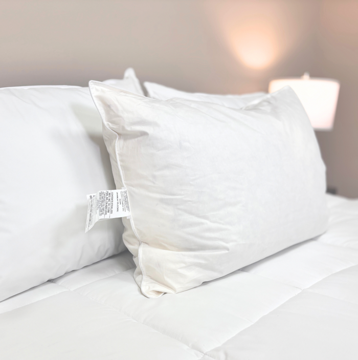 A Pillow Factory Downlite Down Halo pillow with a visible care tag rests on a neatly made bed with white bedding. Soft lighting and cozy beige walls enhance the inviting, hotel-quality bedroom atmosphere.