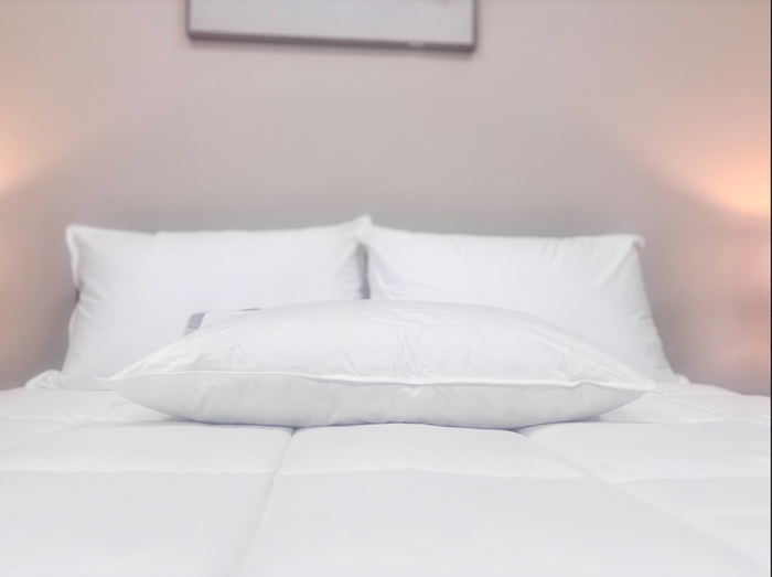 A neatly made bed with three large Pillow Factory Downlite Down Halo pillows, each with a three-chamber design. Warm side lighting and a blurred frame above create a hotel-preferred quality atmosphere.