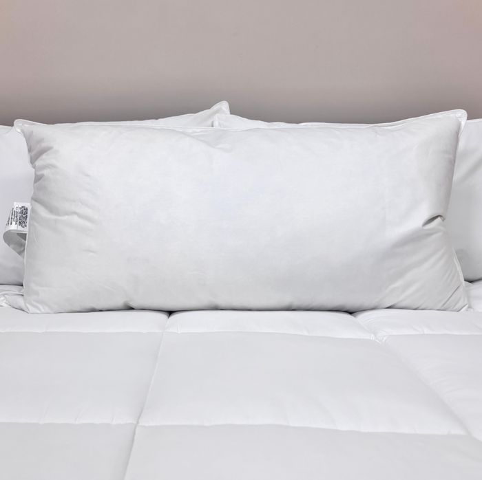 A Pillow Factory Downlite Premium Down Halo pillow sits on a white quilted bedspread, with another pillow partly visible behind it. A plain, light wall highlights the clean, minimal look of the bedroom.