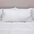 A Pillow Factory Downlite Premium Down Halo pillow sits on a white quilted bedspread, with another pillow partly visible behind it. A plain, light wall highlights the clean, minimal look of the bedroom.