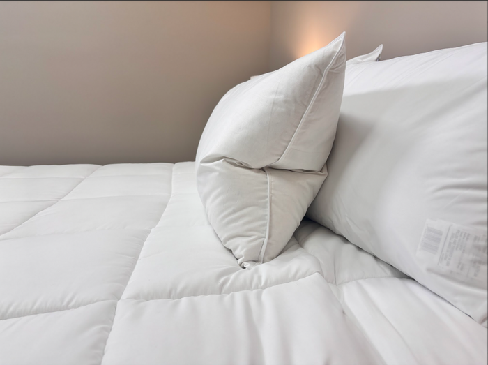 A close-up of a neatly made bed features two Pillow Factory Downlite Premium Down Halo pillows stacked against the headboard and a quilted white comforter. The minimal, clean scene is softly lit, showcasing hotel-quality bedding.