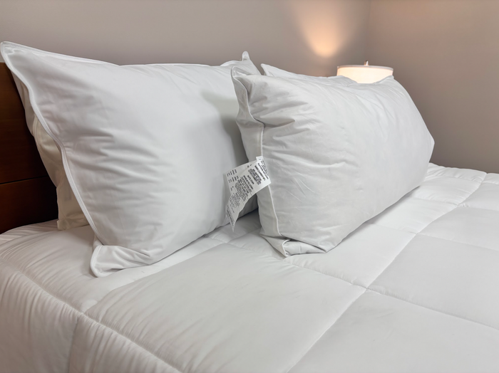 Two large, white Pillow Factory Downlite Premium Down Halo pillows with visible tags rest upright against a wooden headboard on a neatly made bed with a white comforter, while a glowing lamp adds a cozy, hotel-quality touch.