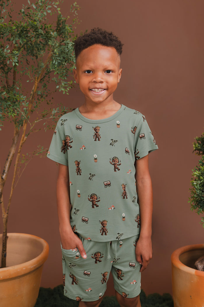 Boy smiling in Green Drawstring Short in Groot print under a similar print Toddler Basic Tee with plant pots around