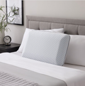 A modern bedroom featuring a gray upholstered headboard, white bedding, and the Malouf EverRest Bamboo Charcoal Pillow with Cooling Cover in a blue-and-white geometric pattern. A black nightstand holds a clock, potted plant, and wall art hangs above.
