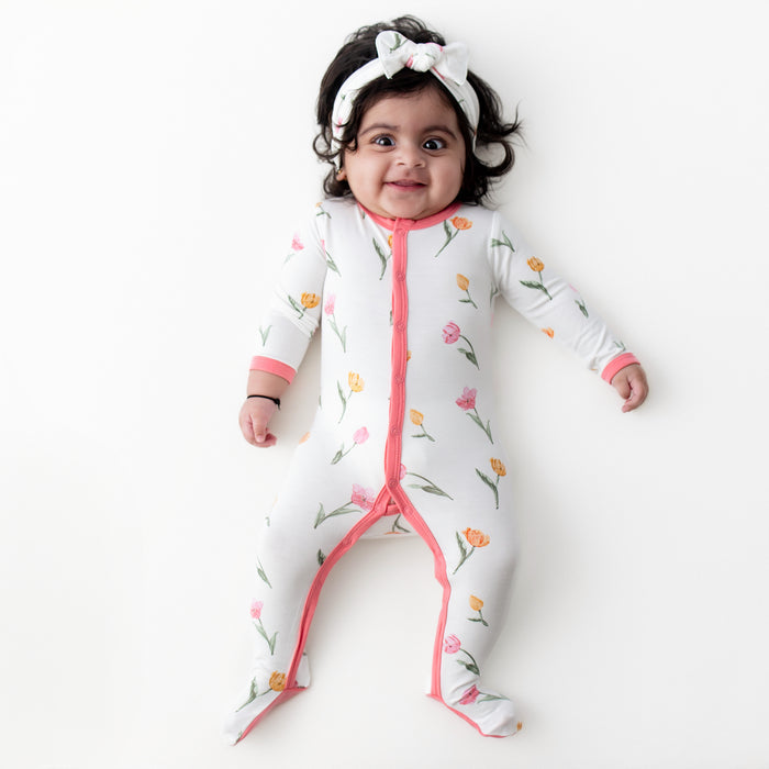 Young infant laying on a white surface wearing the Footie in Tulip with matching knotted bow headband