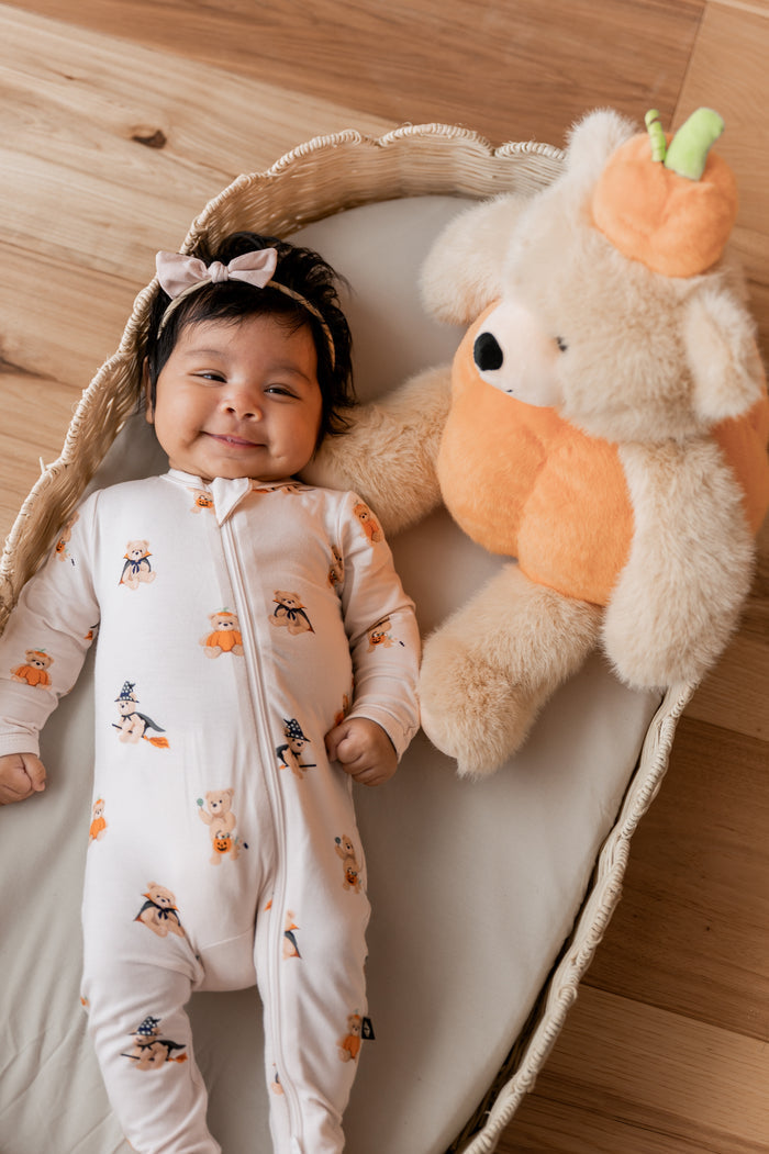 Baby wearing a halloween printed footie with a plush teddy bear dressed as a pumpkin next to her