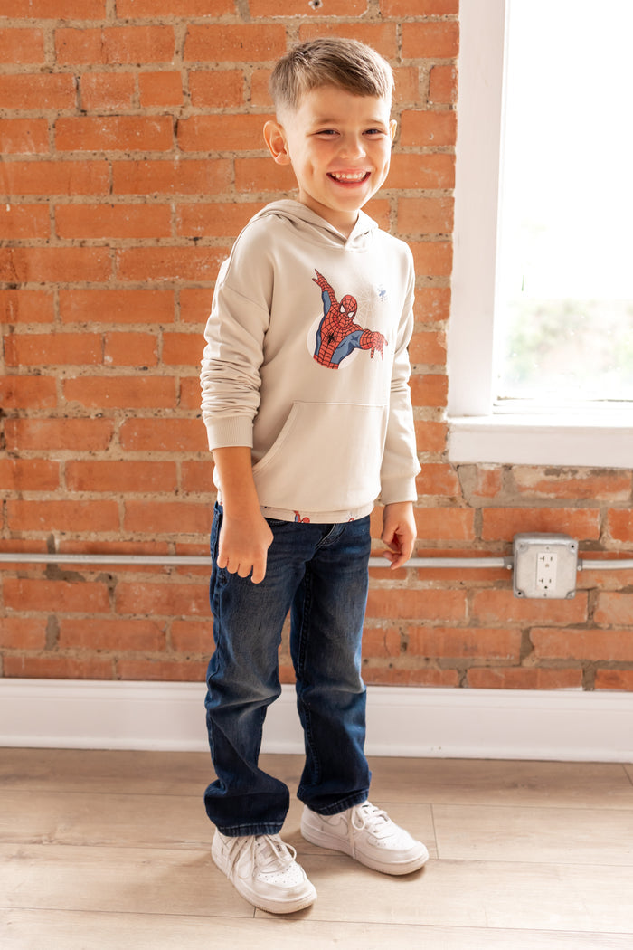 Boy smiling in front of a brick wall wearing an oat color Hoodie with a Spider-Man print in center over a blue jeans and white sneakers