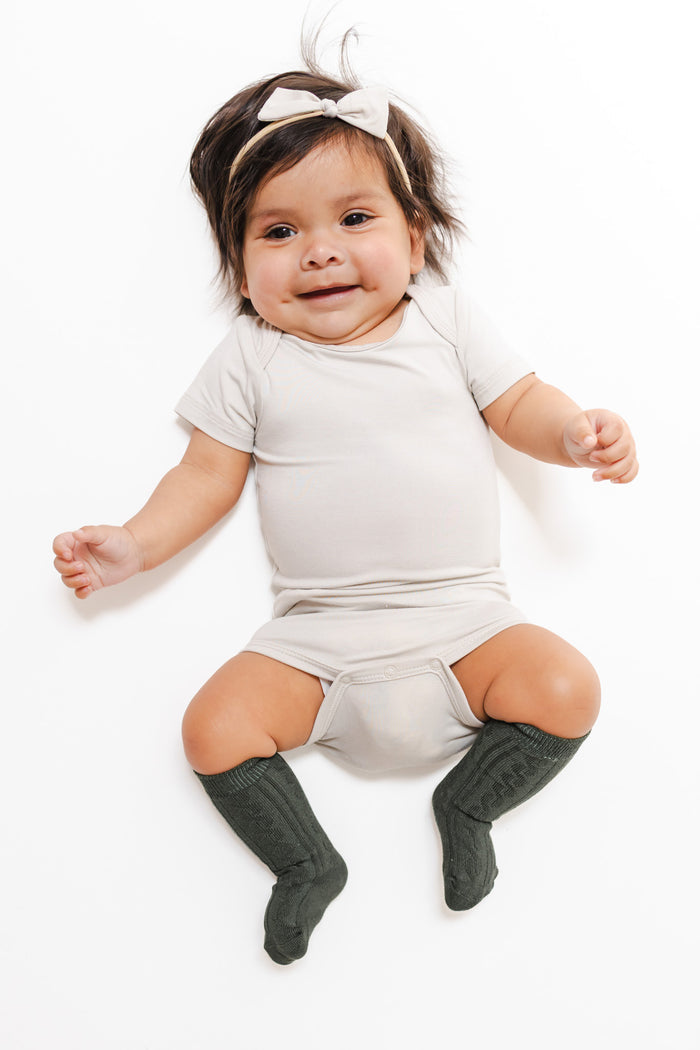 A baby lying on a white background wearing a light beige short-sleeve romper, dark green knee-high socks, and a matching beige bow headband.