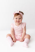 A smiling toddler girl sitting on a white studio floor, wearing a light pink ruffle-sleeve dress, matching pink knee-high socks, and a pink bow headband.