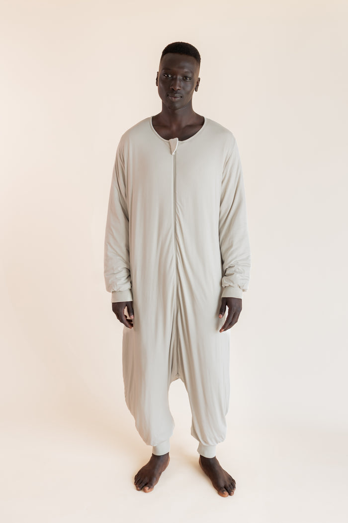A man wearing a light beige, full-body zip-up harem sleep sack against a solid, warm off-white background.