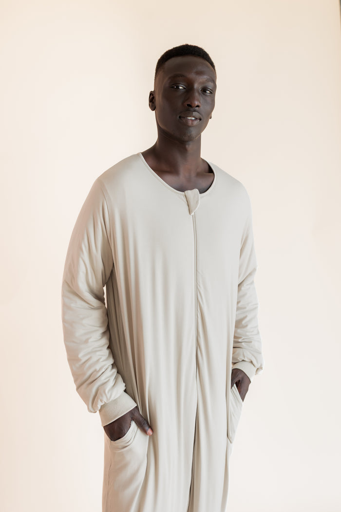 A man wearing a long-sleeved, beige sleep sack with a split neckline and side pockets, standing against a solid light beige background.