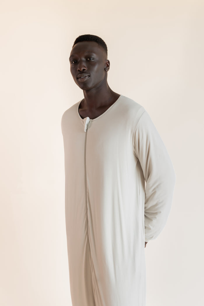 A man wearing a long-sleeved, full-length cream-colored sleep sack , standing against a solid light beige background.