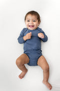 A smiling baby lying on a white background, wearing a long-sleeve blue bodysuit.