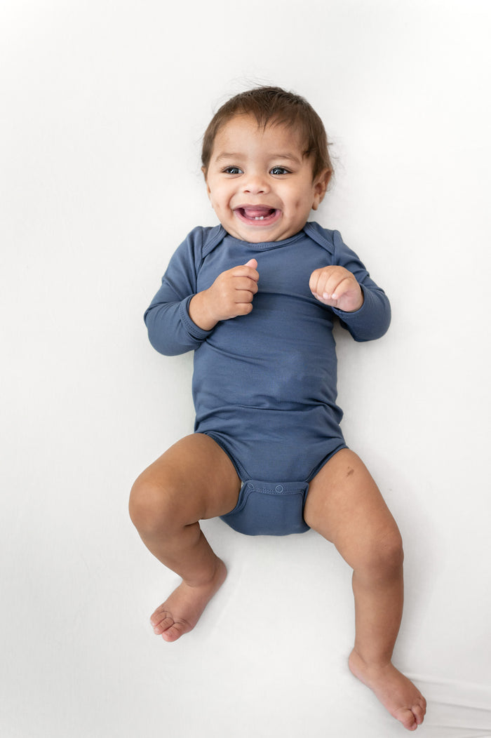 A smiling baby lying on a white background, wearing a long-sleeve blue bodysuit.