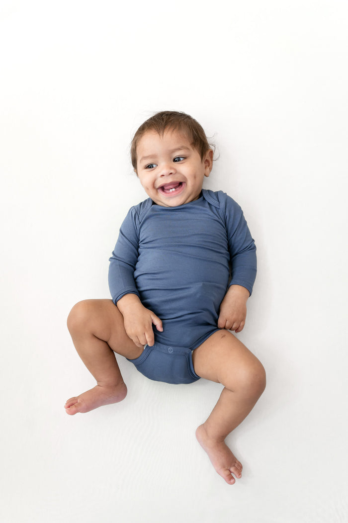 A smiling baby sitting on a white background, wearing a dusty slate blue long-sleeve bodysuit.