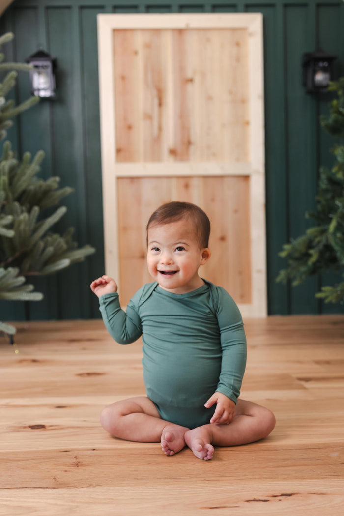 A smiling baby sitting cross-legged on a wooden floor, wearing a long-sleeve sage green bodysuit. The background features a rustic wooden door set against a dark green paneled wall, with decorative lights and greenery on either side.