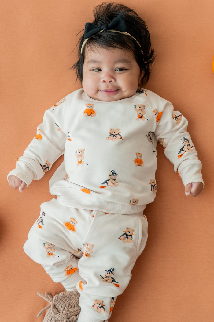 A baby lying on a peach background with small orange pumpkins around them, wearing an off-white long-sleeve pajama set with a Halloween print of ghosts, witches, and pumpkins. The baby is also wearing a black headband and knit socks.
