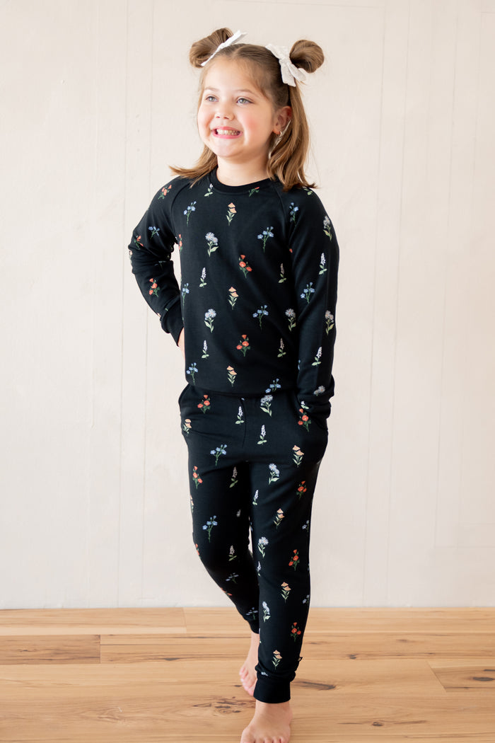 A young girl with brown pigtails wearing a matching black long-sleeve pajama set with a small multicolor winter pattern, standing barefoot on a wooden floor against a white paneled wall.