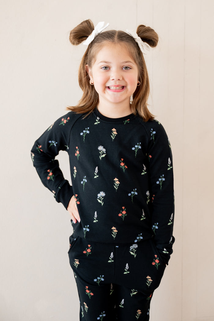 A young girl with brown hair in two buns wearing a black long-sleeve pajama set with a small multicolor floral print, standing against a light beige wall.